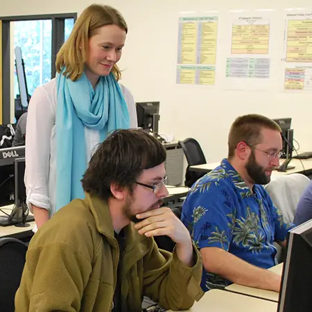 Computer science instructor stands over the shoulders of two students working on a project.