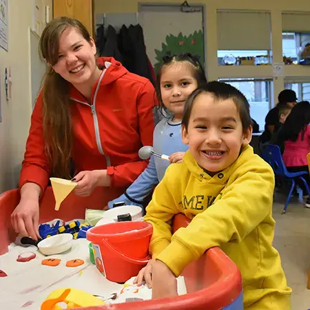 Two young children and a smiling caregiver play with toys in an indoor sandbox.