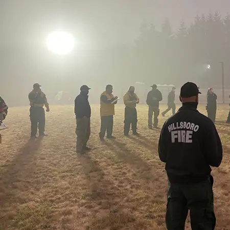 A diverse group of emergency management personnel — ranging from firefighters to EMTs to incident managers — standing in an open field during wildfires making emergency coordination plans.